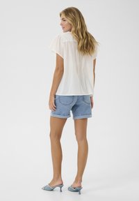 Blouse blanche à manches courtes associée à un short en denim bleu clair, présentant une coupe décontractée et des ourlets retroussés. Des mules à talons bleus complètent le look.