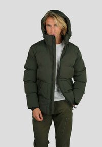 Homme portant une veste à capuche matelassée verte olive et un pantalon assorti avec un t-shirt blanc, se tenant devant un fond gris clair uni.