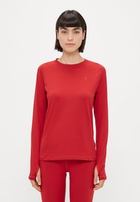 Červená, dlhá športová blúzka s raglánovými rukávmi vyrobená z hladkej, elastickej látky a s malým logom na ľavom hornom rohu.