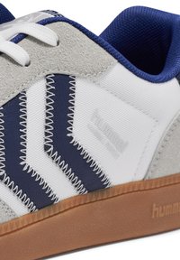 Gros plan sur une basket gris, blanc et bleu marine avec des lacets blancs, un motif chevron cousu, un rembourrage intérieur bleu et une semelle en caoutchouc marron.