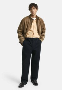 Braune Corduroy-Bomberjacke, hellorange gestreiftes Hemd, marineblaue Hose und dunkle Schuhe. Einfaches Design mit lockerer Passform und minimalen Details.
