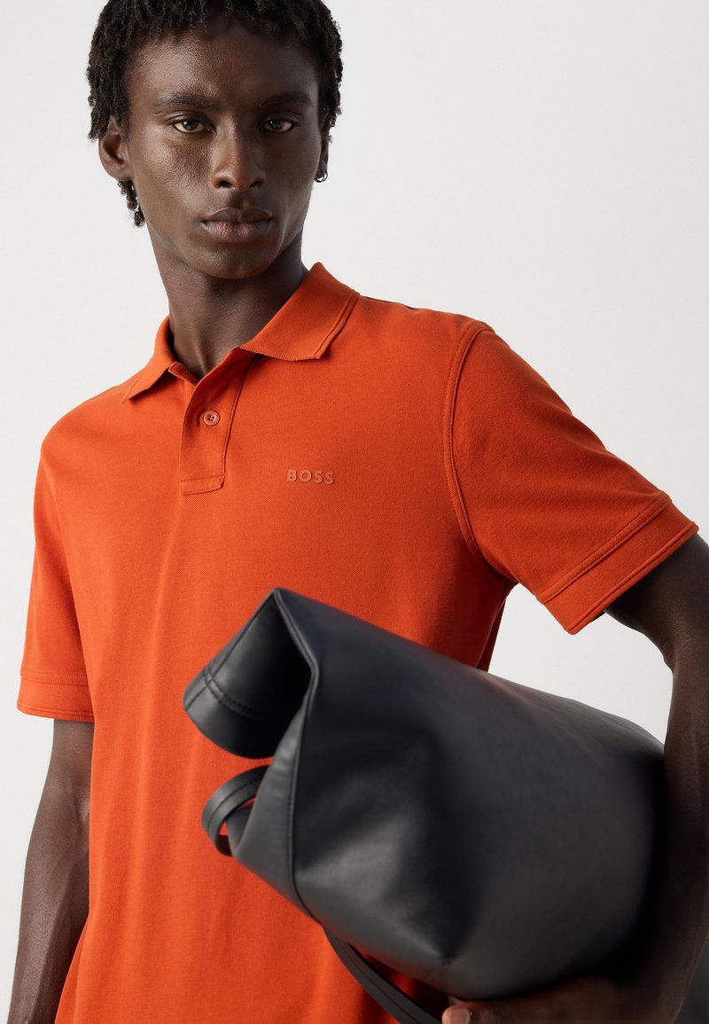 Jeune homme portant un polo orange, tenant un sac en cuir noir enroulé contre un fond clair uni.