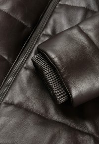 Giacca puffer in cuoio marrone con un design trapuntato, finitura liscia e dettagli in maglia a coste sui polsini. Chiusura frontale con zip visibile.