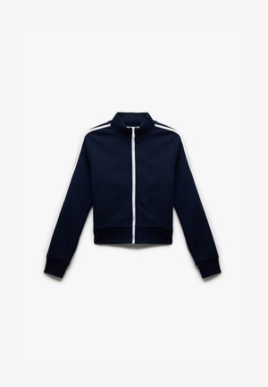 Tummansininen vetoketjullinen urheilutakki, jossa on valkoiset sivuraitat. Ominaisuuksiin kuuluu korkea kaulus ja ribattu hihansuu. Valmistettu sileästä, joustavasta kankaasta.