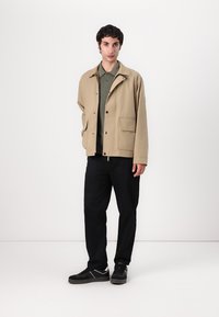 Beige lightweight jacket with a button-down front, two front pockets, and a collar, worn over a green ribbed sweater and black pants.