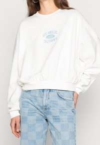 Bílá mikina s kulatým výstřihem, volným střihem, elastickým pasem a světle modrým potiskem "Los Angeles 1992 California" přes hruď.