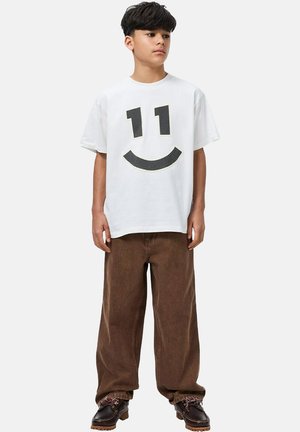 Wit katoenen T-shirt met een grafische zwarte mesh van "11" en een glimlach; gecombineerd met bruine wijde broekspijpen en donkere schoenen.