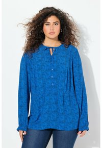 Blaue Langarmbluse mit wellenförmigem schwarzen Muster, Schlüsselaugen-Ausschnitt, fünf Knöpfen und einer Vorderasche, kombiniert mit dunklen Jeans.