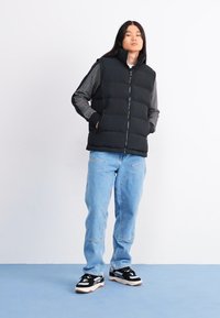 Gilet noir matelassé avec col haut sur une chemise grise à manches longues, associé à un jean bleu clair et des baskets noires. Design simple, sans motifs visibles.