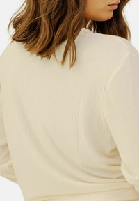 Top côtelé beige clair avec une coupe décontractée, présentant un col rond et une texture de tissu douce, partiellement montré de l'arrière.