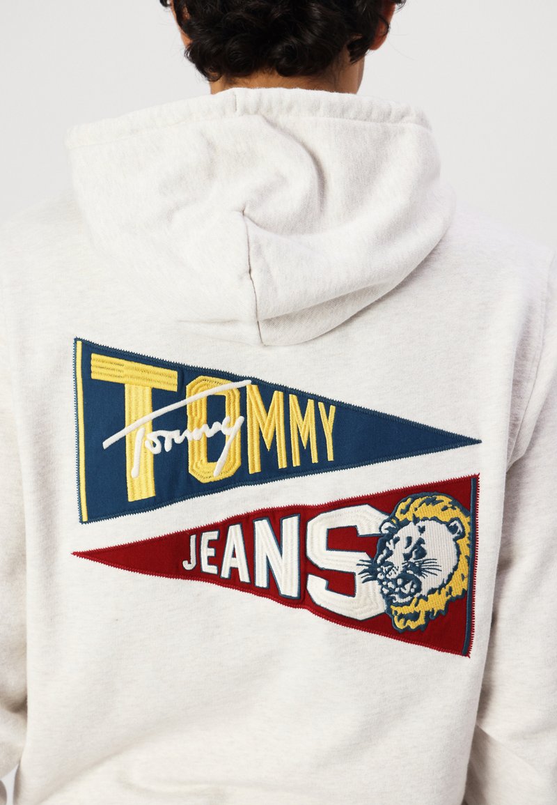 Lichtgrijze hoodie met een groot geborduurd ontwerp met blauwe en rode vlaggen, de tekst "TOMMY" in het geel, en een gedetailleerde leeuwgrafiek.
