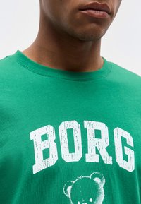 Κοντινή λήψη ενός ατόμου που φοράει πράσινο πουκάμισο με λευκά μπλοκ γράμματα σε φθαρμένο στυλ που γράφουν «BORG» και ξεθωριασμένο γραφικό με αρκουδάκι από κάτω.