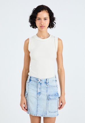 Lichtbeige mouwloze top gecombineerd met een vervaagde blauwe denimrok met een zak en naaddetails. Zachte textuur over het geheel.