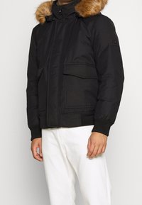 Schwarze gepolsterte Jacke mit einer abnehmbaren braunen Fellkapuze, Frontreißverschluss, zwei großen Taschen und gerippten Bündchen. Das Material wirkt glatt mit einer matten Oberfläche.