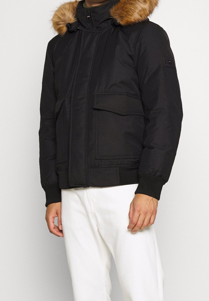 Schwarze gepolsterte Jacke mit einer abnehmbaren braunen Fellkapuze, Frontreißverschluss, zwei großen Taschen und gerippten Bündchen. Das Material wirkt glatt mit einer matten Oberfläche.