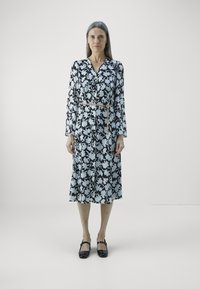 Langärmliges Kleid mit einem schwarz-weißen Blumenmuster, einer taillierten Taille, die durch einen goldenen Kettengürtel betont wird. Die Schuhe sind schwarze Ballerinas.