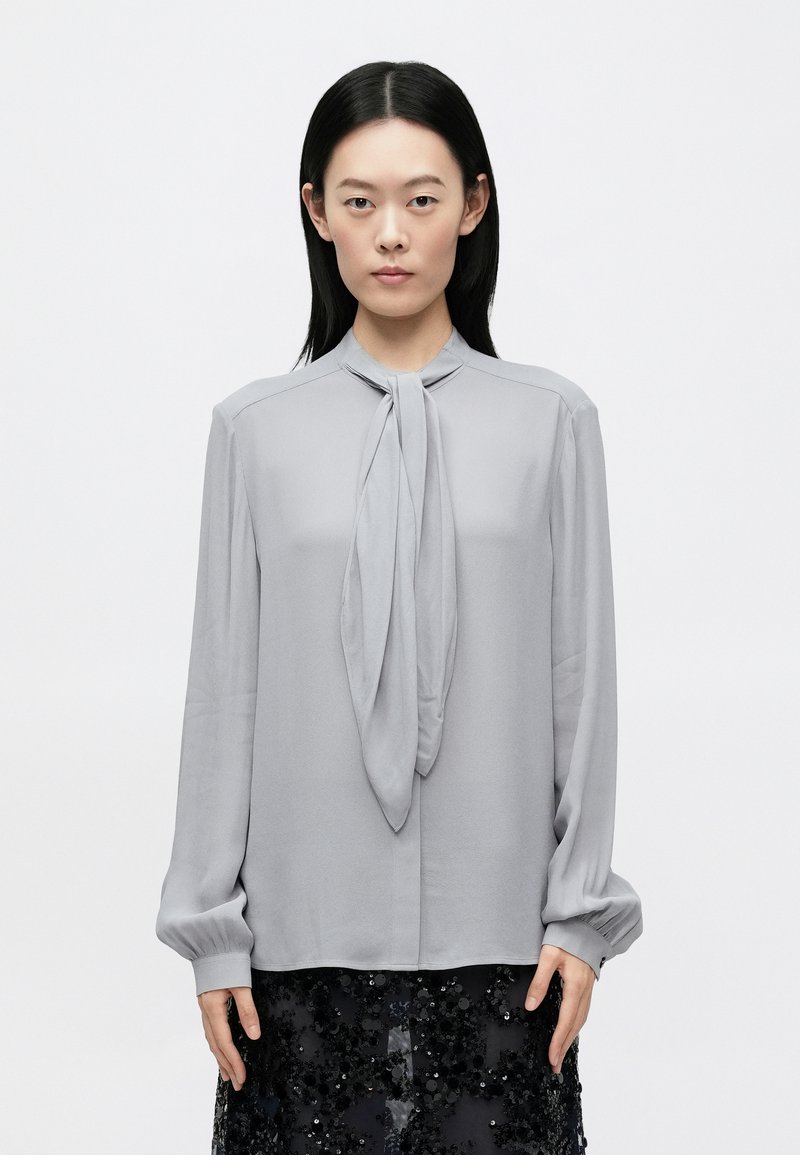 Jasnoszara bluzka z gładkiego materiału, z wiązanym dekoltem, długimi rękawami ze ściągaczami i prostym krojem.