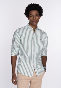 Camisa de botones a rayas en verde claro y blanco. Presenta un cuello alto, mangas largas arremangadas hasta los codos y un detalle sutil del logo.