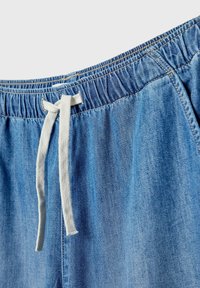 Pantalon en denim bleu avec taille élastique et cordon blanc noué en nœud sur un fond uni.