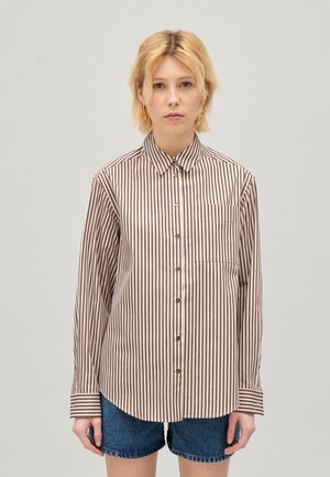 Młoda osoba z krótkimi blond włosami, nosząca złote kolczyki w kształcie kół, brązową i białą pionową w paski koszulę na guziki oraz dżinsowe szorty.