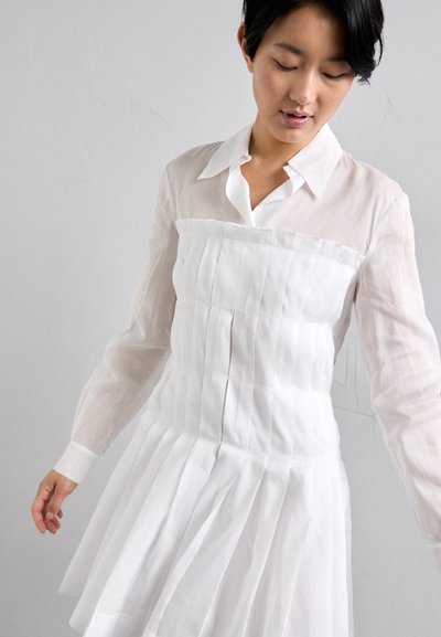 Abito bianco plissettato con collo a camicia e maniche lunghe e trasparenti. Il tessuto appare leggero e texture, creando un effetto a strati.