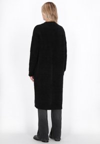 Cardigan lung negru pufos, cu o croială lejeră, având guler rotund, mâneci lungi și fante laterale. Împreună cu pantaloni gri închis și botine negre.