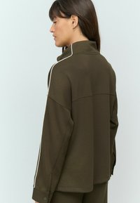 Veste marron surdimensionnée avec des rayures blanches sur les côtés, col haut et texture douce ; possède des poignets à boutons et une coupe décontractée.
