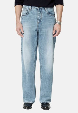 Man draagt lichtblauwe wijde jeans, een zwart shirt met lange mouwen, zilveren armbanden en zwarte schoenen, staand tegen een effen achtergrond.