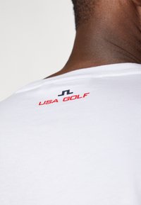T-shirt de algodão branca com decote redondo, com "USA GOLF" em vermelho e um logótipo preto na parte de trás da gola. Textura suave.
