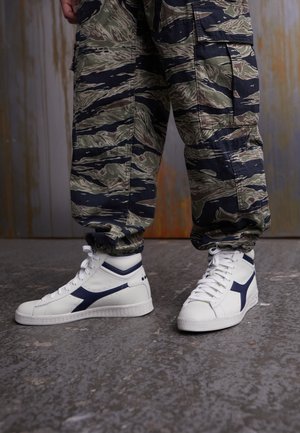 Unterschenkel einer Person, die Camouflage-Cargohosen und weiße High-Top-Sneakers mit schwarzen Akzenten trägt und auf einem strukturierten Betonboden steht.