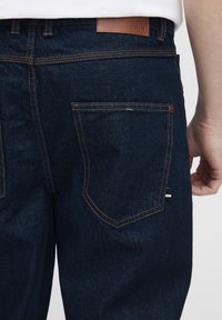 Jeans de mezclilla azul oscuro con costuras naranjas, que presentan una etiqueta de cuero en la cintura y un bolsillo trasero con una etiqueta a rayas.