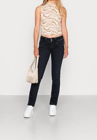 Kvinna klädd i en ärmlös zebra-mönstrad cropped topp, svarta jeans, vita plattformssneakers, hållande en beige handväska vid sin sida.