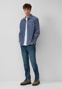 Blauwe corduroy overhemd met knopen en een borstzak, gedragen over een wit T-shirt, gecombineerd met donkerblauwe jeans en bruine veterschoenen.