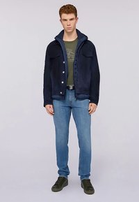 Tmavě modrá fleecová bunda s kapucí, zapínáním na knoflíky, dvěma kapsami na prsou, v kombinaci se zeleným tričkem a světle modrými džínami, nošena s tmavými teniskami.