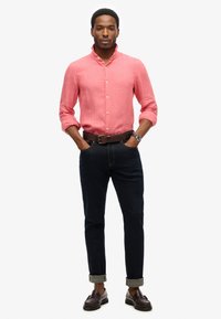 Lichtroze linnen knoopshirt met een bandkraag, gecombineerd met donkerblauwe jeans en een paar bruine loafers. Opgerolde manchetten op de jeans.