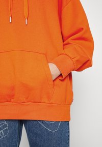 Sudadera naranja hecha de tela suave con un bolsillo canguro y puños acanalados, combinada con unos vaqueros azules que presentan un detalle sutil.