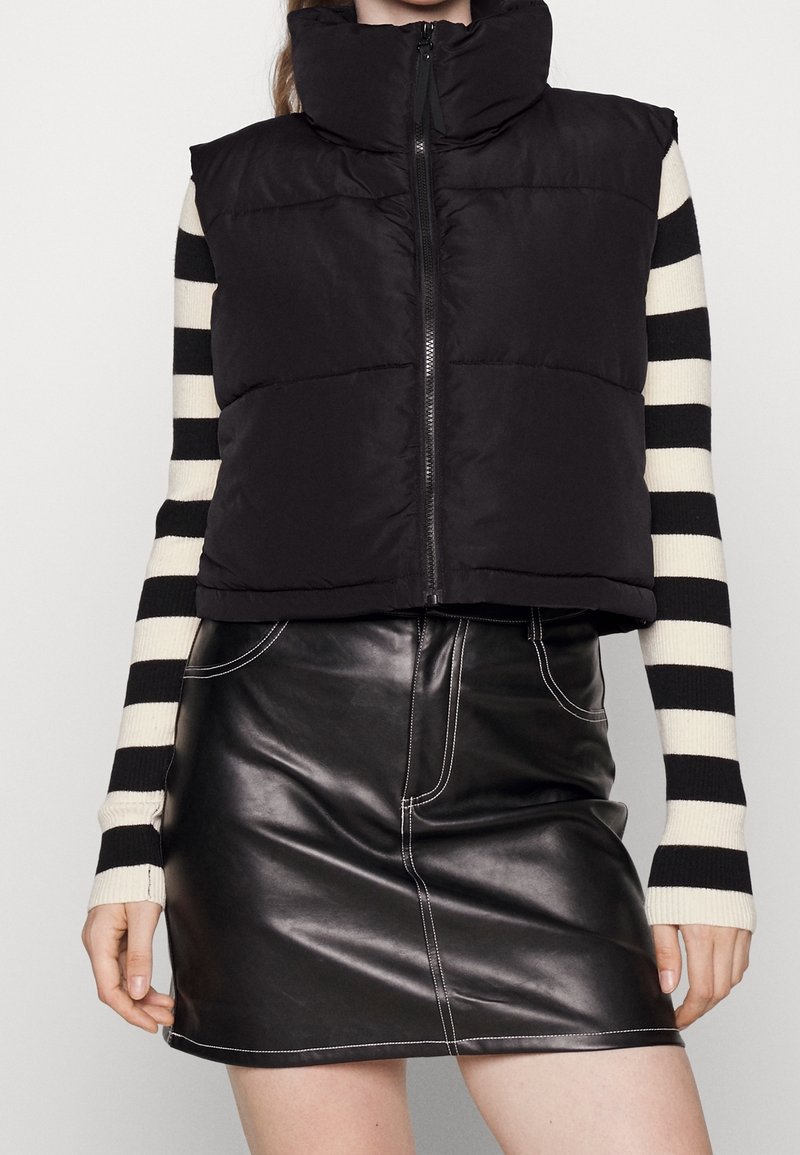 Czarna kamizelka puchowa z wysokim kołnierzem i zapięciem na zamek, noszona na czarnej i białej w paski bluzce z długim rękawem, zestawiona z czarną skórzaną spódnicą.