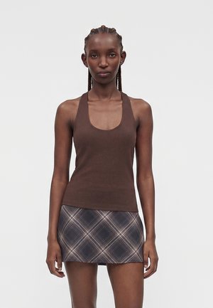 Kobieta z warkoczami, ubrana w brązowy top bez rękawów i czarno-białą kratkę mini spódniczkę, stojąca na jednolitym tle.