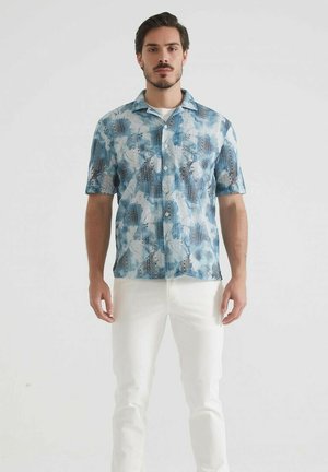 Chemise à manches courtes à boutons dans des tons de bleu avec des motifs abstraits ; le tissu semble léger avec une coupe décontractée, associée à un pantalon blanc.