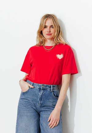 Femme blonde aux cheveux lâchés portant un t-shirt rouge à imprimé cœur, rentré dans un jean bleu taille haute, debout devant un fond blanc.