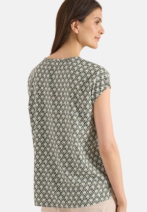 Nainen lyhythihaisessa, vihreävalkoisella geometrisella kuviolla varustetussa paidassa, kuvattuna takaa ja sivulta neutraalia taustaa vasten.