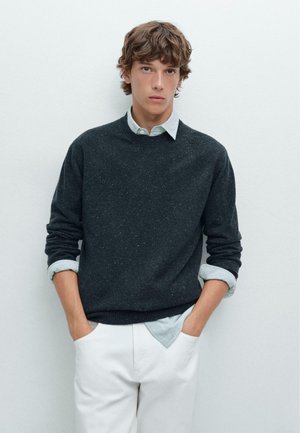 Dark gray speckled knit sweater with a round neckline, layered over a light blue collared shirt, paired with white pants.