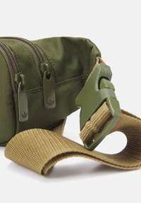 Olivgrön tygpåse med två dragkedjor och märkta dragflikar; har ett beige justerbart band och plastspänne.