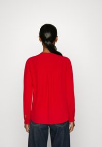 Rote Bluse mit langen Ärmeln und lockerer Passform, mit geradem Saum und Knopfdetails an den Manschetten. Ein Rückenfalte sorgt für zusätzliche Details.