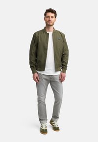 Oliven grøn bomberjakke, hvid t-shirt, lysegrå jeans og oliven sneakers. Har lynlås, ribbede manchetter og sidelommer som detaljer.
