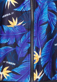 Vêtement noir avec des feuilles tropicales bleues et des fleurs jaunes, fermeture éclair au centre, texte blanc "NEBBIA" sur le tissu.