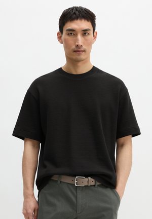 Man draagt een zwart T-shirt met korte mouwen en ronde hals, grijze broek met een beige riem, staande tegen een effen witte achtergrond.