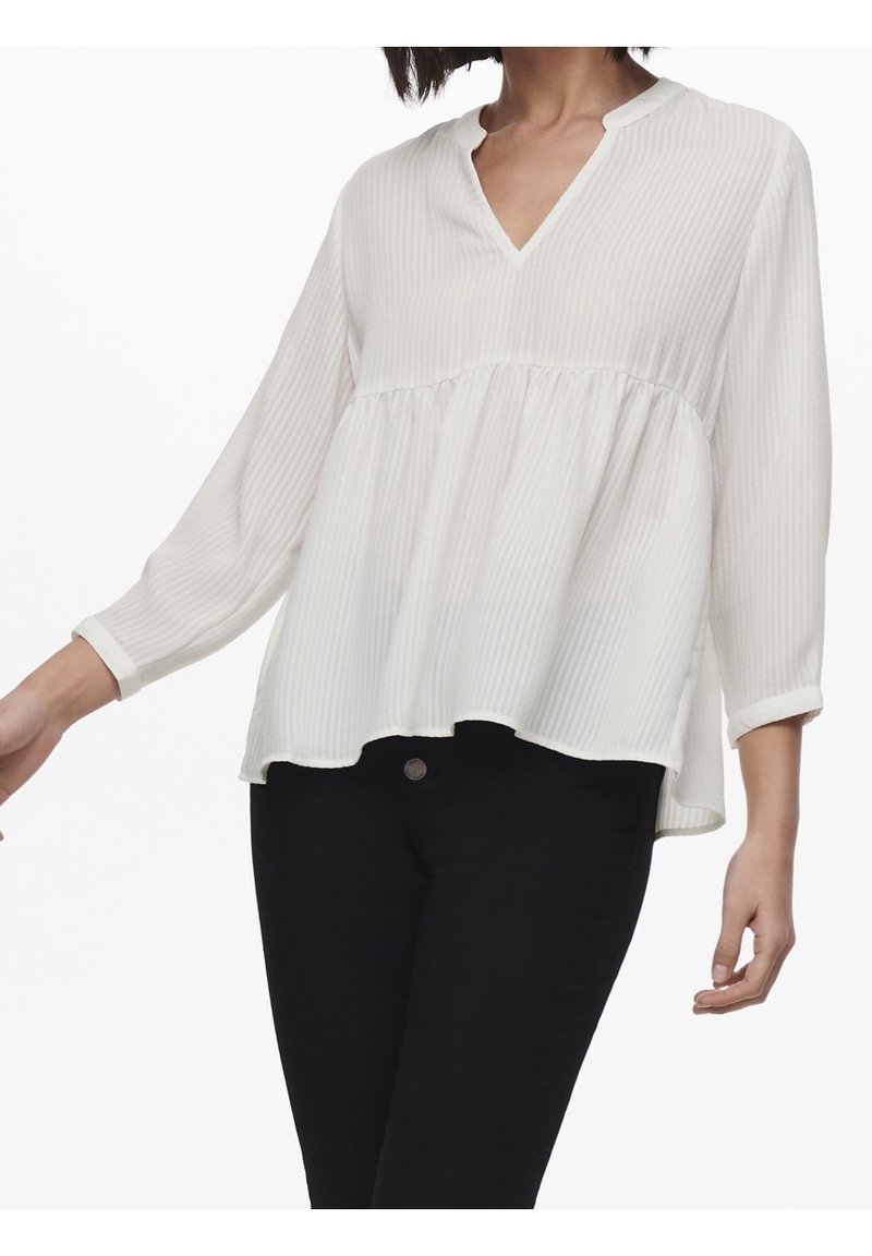 Blouse blanche à manches longues avec un col en V, présentant une coupe ample et une texture côtelée horizontale. Assortie à un pantalon noir.