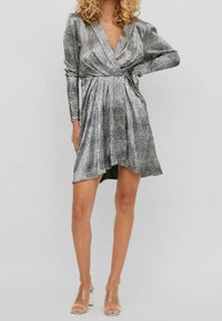 Robe drapée métallique argentée avec des manches longues, décolleté en V, taille cintrée et jupe fluide avec un ourlet asymétrique. Sandales à talons transparents.