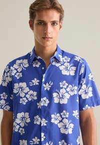 Camisa de manga corta de tela azul con grandes flores de hibisco blancas, frente abotonado y cuello clásico. Textura suave.